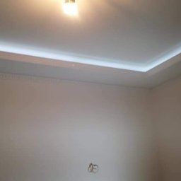 SLAVLEN GRUP Viacheslav Kudinov - Wykończony sufit podwieszany z oświetleniem LED i dekoracyjnym gzymsem, białe ściany w pomieszczeniu przed umeblowaniem.