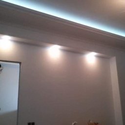 SLAVLEN GRUP Viacheslav Kudinov - Mężczyzna maluje ścianę w pokoju z podwieszanym sufitem i oświetleniem LED, stojąc na drewnianym podeście.