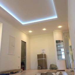 SLAVLEN GRUP Viacheslav Kudinov - Pokój w trakcie remontu: widoczna podwieszana płyta karton gips z oświetleniem LED, świeżo pomalowane ściany, częściowo ułożona podłoga, kartony i narzędzia na podłodze, widok z perspektywy osoby...