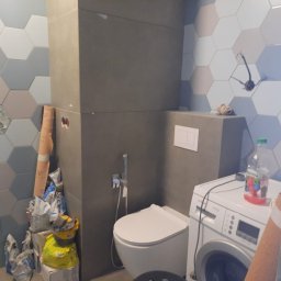 SLAVLEN GRUP Viacheslav Kudinov - Łazienka w trakcie remontu: widoczne płytki heksagonalne na ścianie, zabudowa z szarego betonu architektonicznego, muszla klozetowa, pralka oraz worki z zaprawą i kartonowe pudełka. Niedokończone...