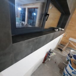 SLAVLEN GRUP Viacheslav Kudinov - Montaż okna z ciemną ramą i parapetem w pomieszczeniu z drewnianą boazerią w Sosnowcu, widoczne narzędzia i wiaderka z farbą.