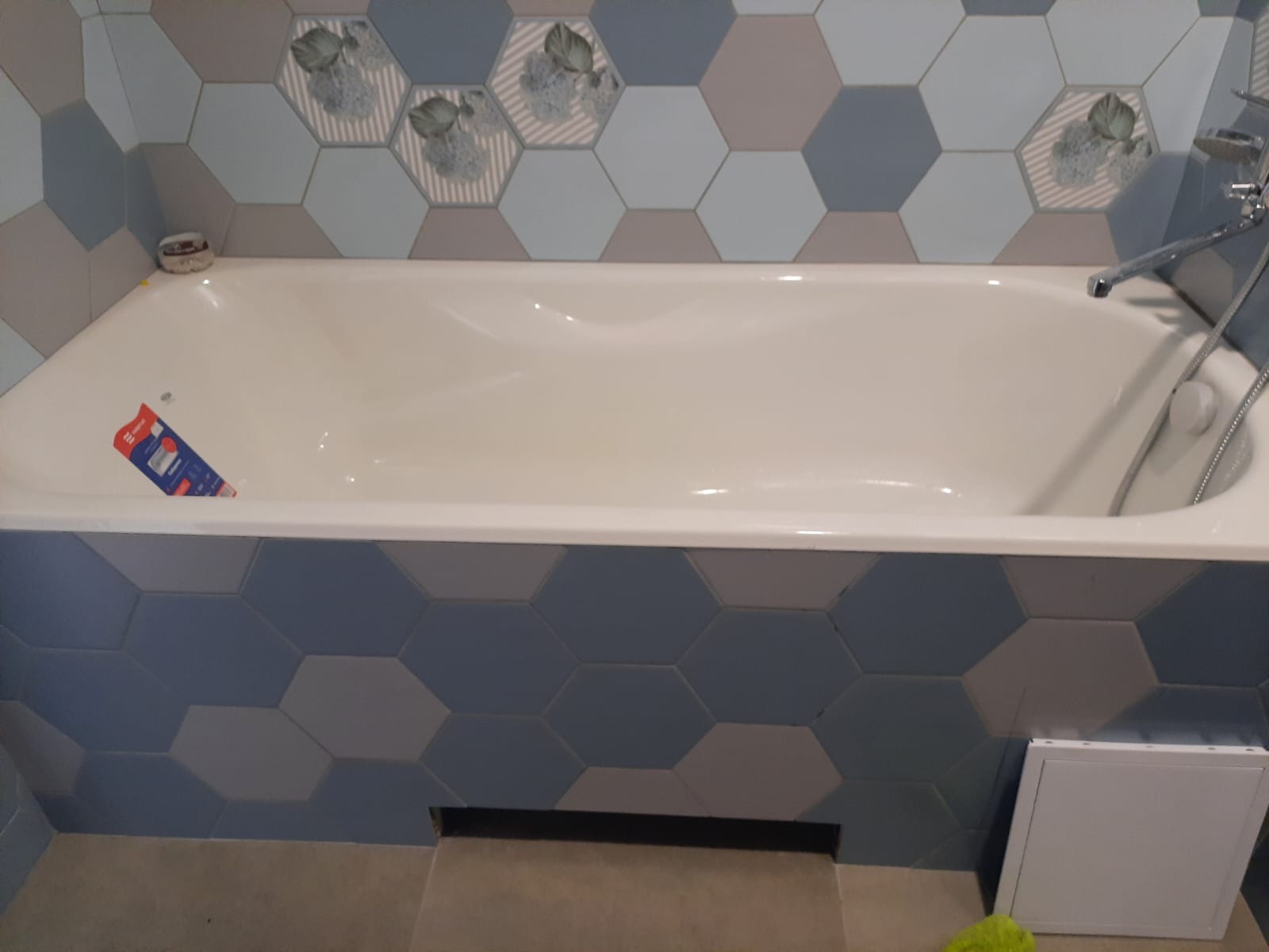 Biała wanna obudowana mozaiką z heksagonów w odcieniach szarości i błękitu, z dekoracyjnymi płytkami z motywem roślinnym na ścianie, widoczny fragment baterii i syfonu.