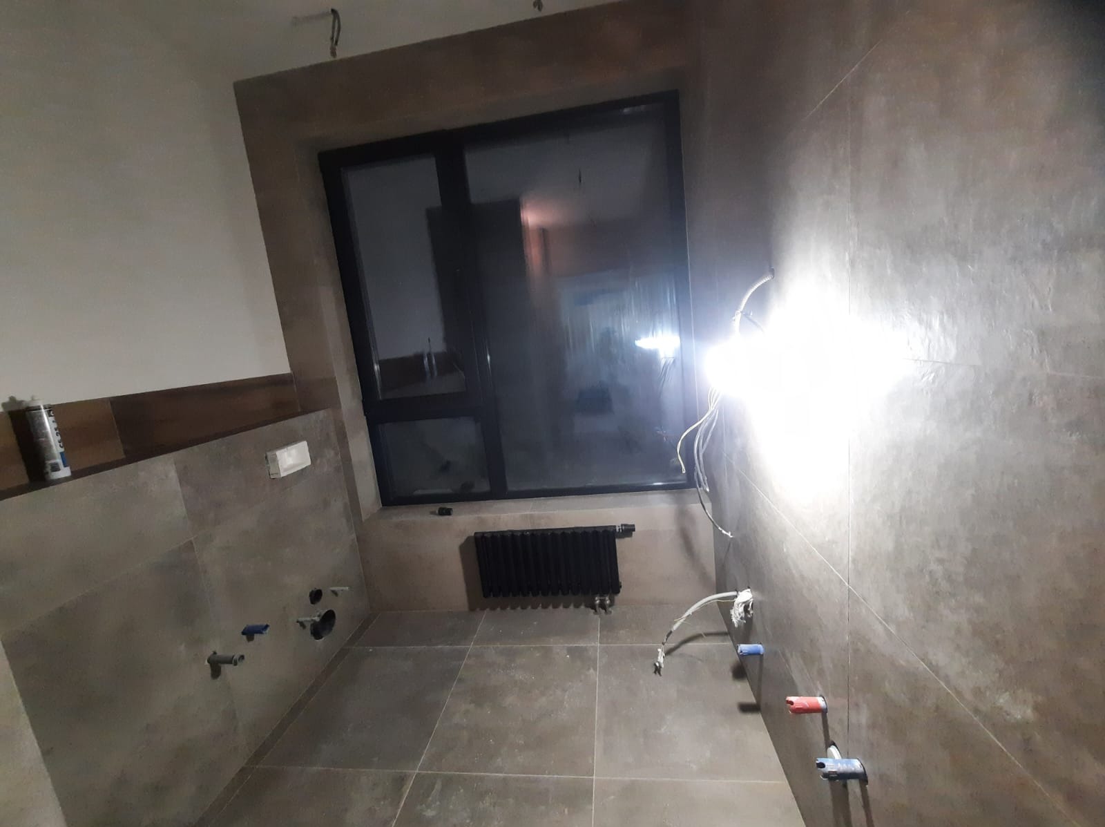 Łazienka w trakcie wykańczania z szarymi płytkami, widoczne wyprowadzenia instalacji wodnej, grzejnik pod oknem i puszka elektryczna, oświetlona mocnym światłem roboczym.