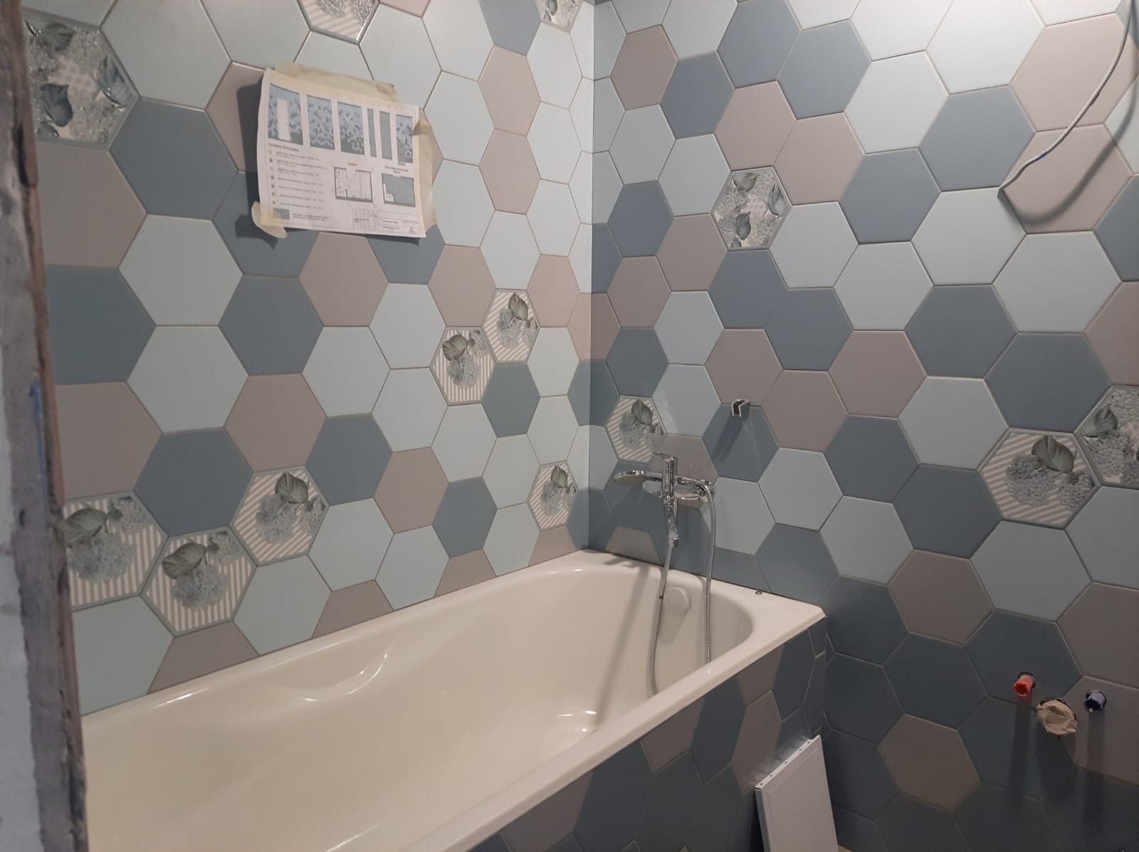 Wanna obłożona heksagonalnymi płytkami w odcieniach szarości i błękitu, niektóre z motywem liści, z widoczną baterią i planem łazienki przyklejonym do ściany.