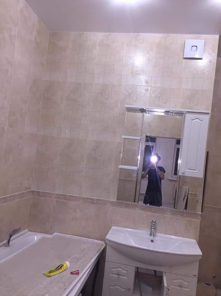 Wykończona łazienka z beżowymi płytkami, białą umywalką z szafką i wanną, widoczne odbicie osoby robiącej zdjęcie w lustrze.