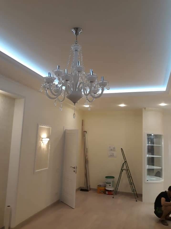 Wykończone wnętrze pokoju z białym sufitem podwieszanym z oświetleniem LED, ozdobnym żyrandolem i ścianami w odcieniach beżu, widoczne drabina malarska i akcesoria malarskie.