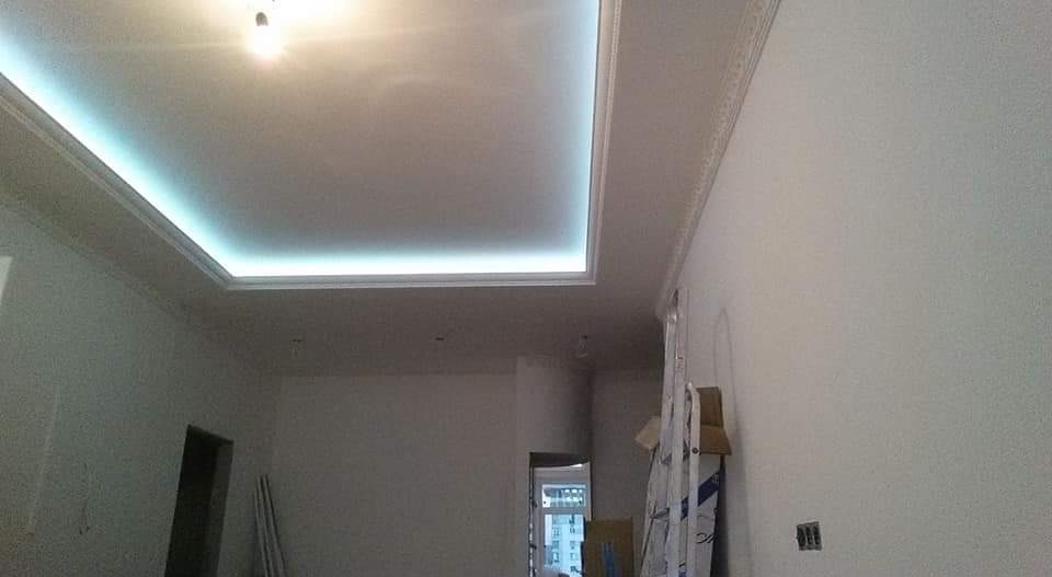 Podwieszany sufit z oświetleniem LED w kształcie prostokąta, listwy dekoracyjne na ścianie i suficie, widoczna drabina i kartony w tle, surowy stan wykończenia wnętrza.