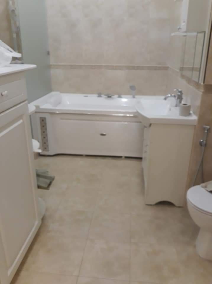 Wykończona łazienka z beżowymi płytkami, białą wanną z hydromasażem i umywalką z szafką, widoczna toaleta i fragment szafki.