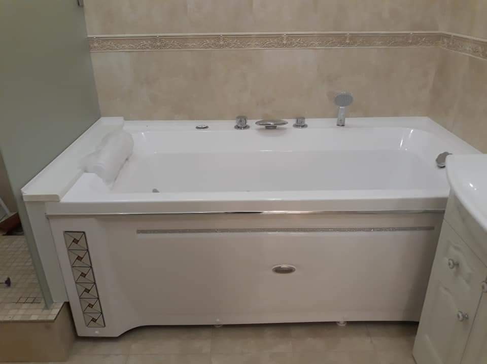 Nowoczesna, biała wanna z hydromasażem w łazience z beżowymi płytkami, widoczna armatura i panel dekoracyjny z geometrycznym wzorem.