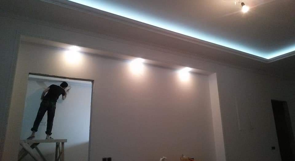 Mężczyzna maluje ścianę w pokoju z podwieszanym sufitem i oświetleniem LED, stojąc na drewnianym podeście.