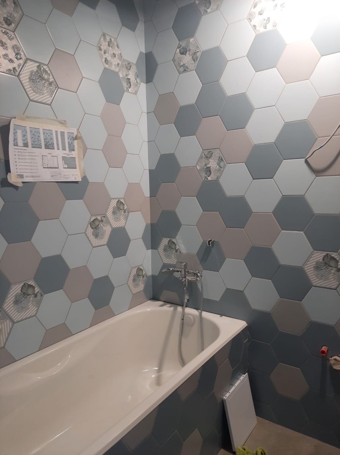 Wykończona łazienka z wanną obudowaną heksagonalnymi płytkami w odcieniach szarości, błękitu i beżu, z dekoracyjnymi wstawkami z motywem liści, widoczny schemat montażu płytek na ścianie.
