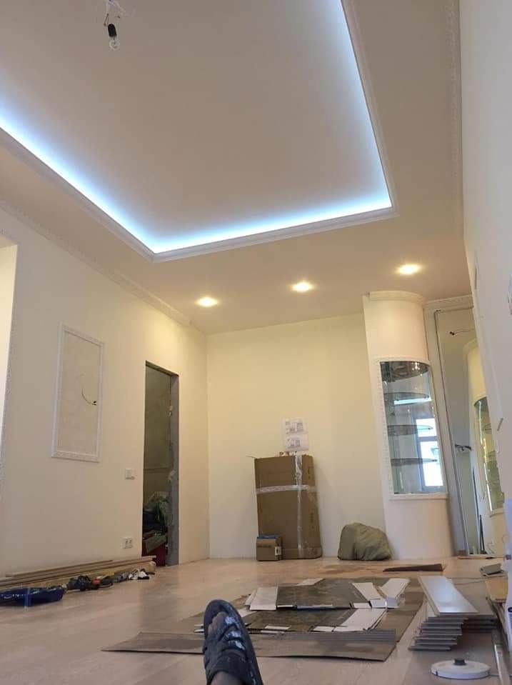 Pokój w trakcie remontu: widoczna podwieszana płyta karton gips z oświetleniem LED, świeżo pomalowane ściany, częściowo ułożona podłoga, kartony i narzędzia na podłodze, widok z perspektywy osoby...