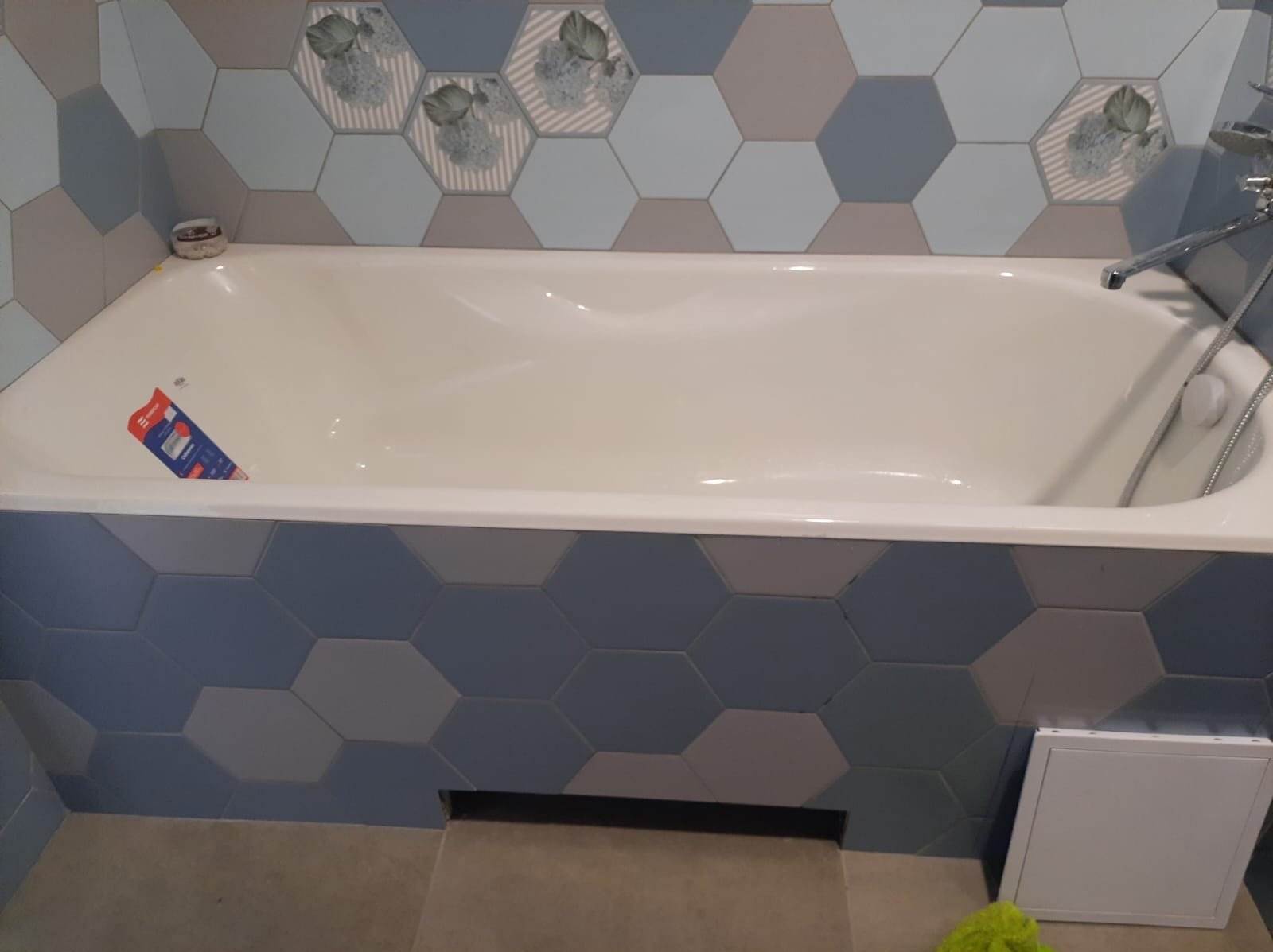 Biała wanna obudowana heksagonalnymi płytkami w odcieniach szarości i błękitu, z widoczną baterią i słuchawką prysznicową, a także fragmentem podłogi i klapą rewizyjną.