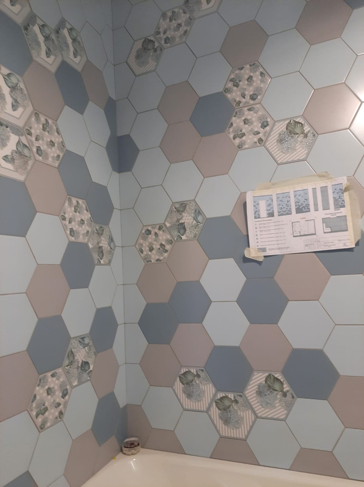 Ściana łazienki wyłożona heksagonalnymi płytkami w odcieniach szarości, błękitu i beżu, niektóre z motywem kwiatowym, z planem łazienki przyklejonym do ściany.