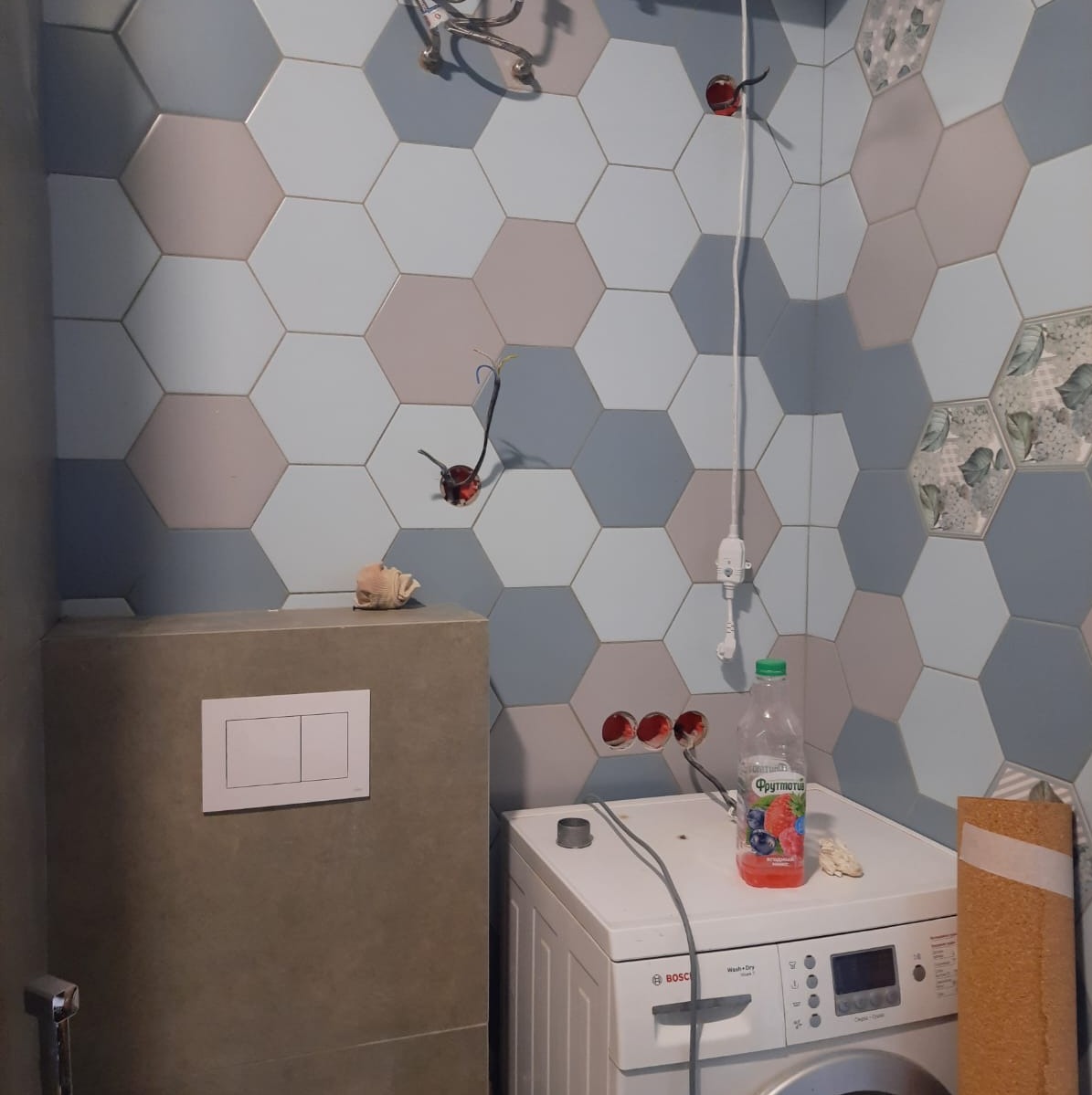 Łazienka w trakcie wykończenia z heksagonalnymi płytkami w odcieniach szarości, błękitu i różu, widoczne niedokończone instalacje elektryczne oraz pralka z butelką soku na górze.