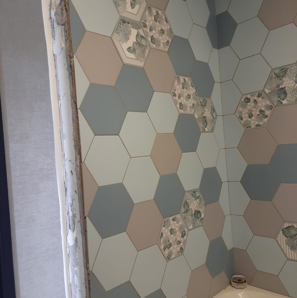 Ściana w łazience wyłożona sześciokątnymi płytkami w odcieniach szarości, beżu i błękitu, z niektórymi płytkami z motywem kwiatowym, widoczny surowy fragment ściany przed wykończeniem.