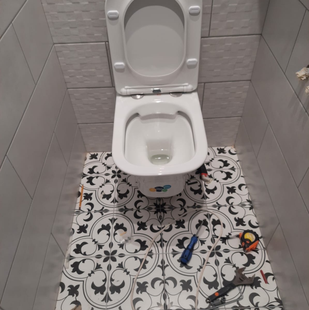 Biała toaleta z otwartą klapą na wzorzystej, czarno-białej podłodze w łazience z szarymi, prostokątnymi płytkami na ścianach, widoczne narzędzia i przewody wskazują na trwający montaż.