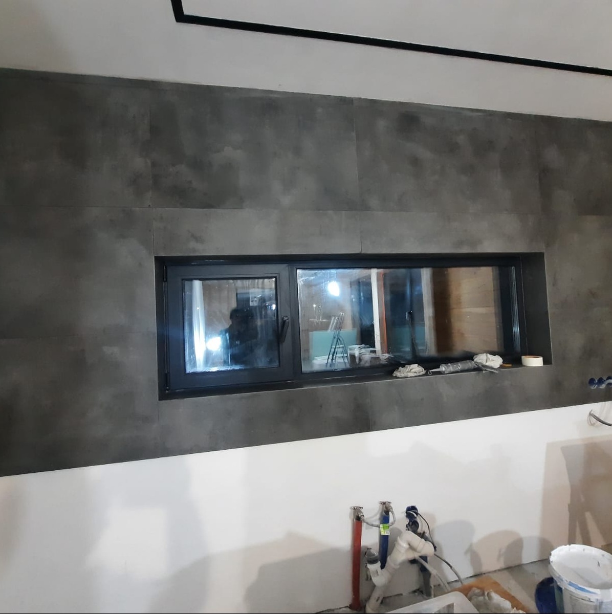 Wykończona ściana w łazience z szarymi płytkami imitującymi beton i wąskim oknem w czarnej ramie; widoczne niezakończone prace instalacyjne poniżej.