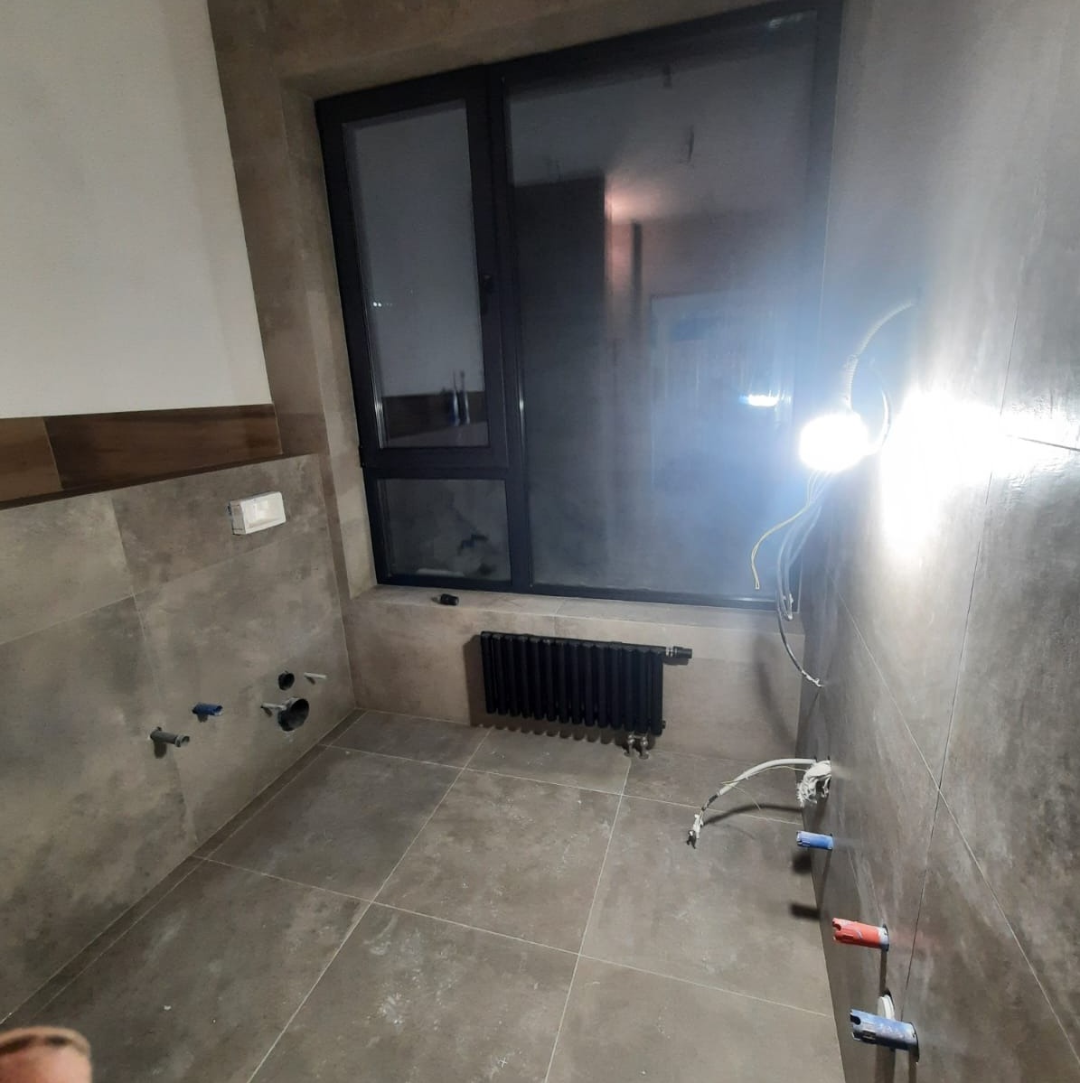 Łazienka w trakcie wykańczania: szare płytki podłogowe i ścienne, czarny grzejnik pod oknem, wyprowadzone instalacje wodne i elektryczne, oświetlenie robocze.