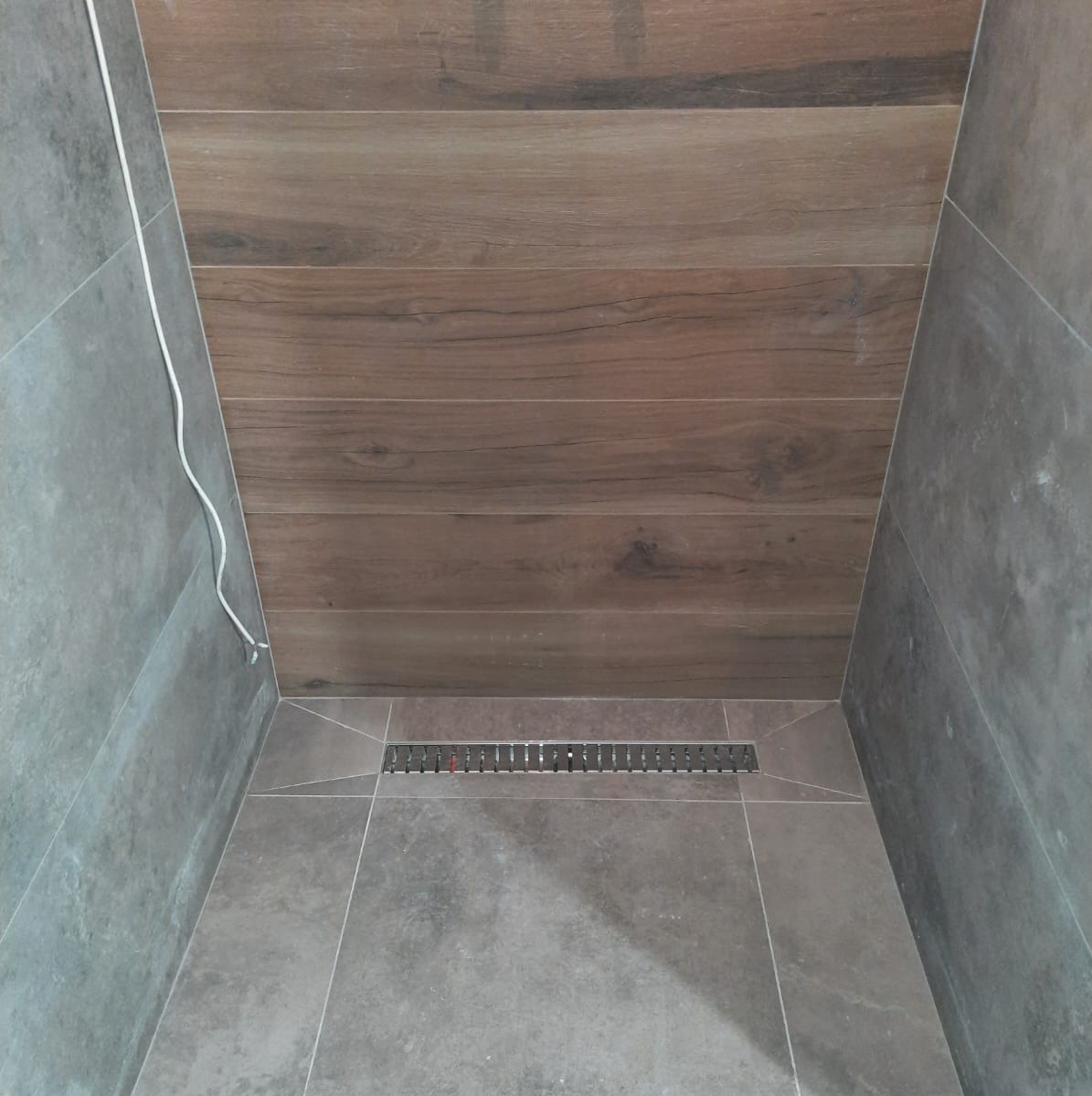 Wykończona kabina prysznicowa z szarymi płytkami i drewnopodobnym sufitem, widoczny odpływ liniowy i wystający przewód elektryczny.