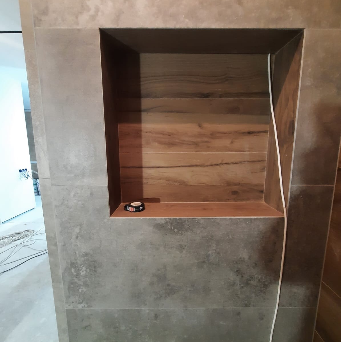 Wnęka w ścianie wyłożona płytkami imitującymi drewno, na półce leży zwinięta miarka, widoczne przewody elektryczne.