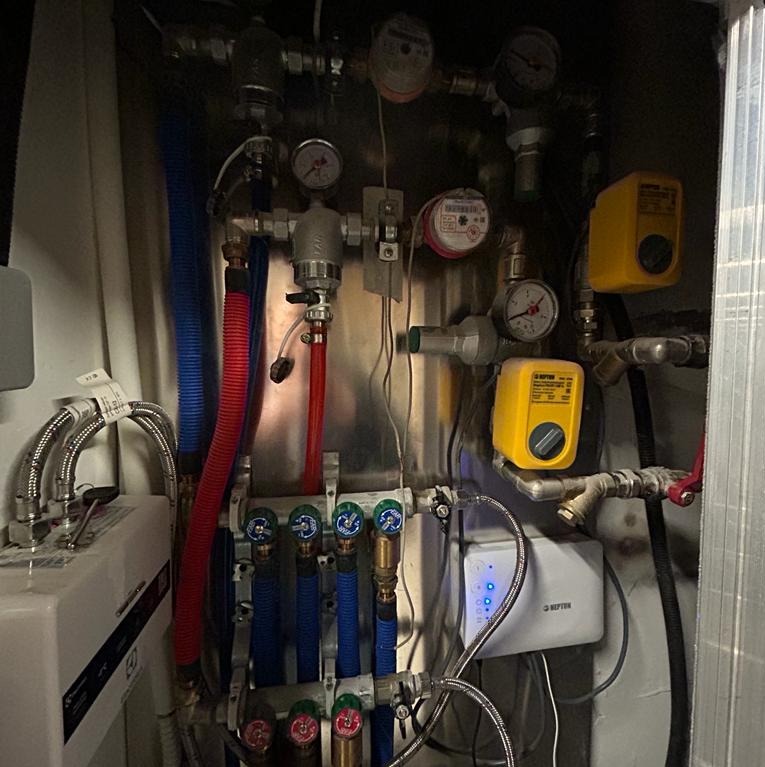 Rozdzielacz ogrzewania podłogowego z rurami w kolorze niebieskim i czerwonym, manometry i regulatory ciśnienia zamontowane na metalowej ścianie.