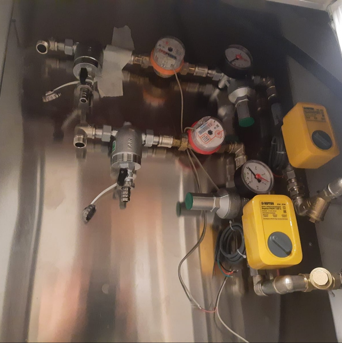Wewnętrzna instalacja wodna z licznikami, manometrami i zaworami odcinającymi, zamontowana na metalowej płycie wewnątrz szafki.