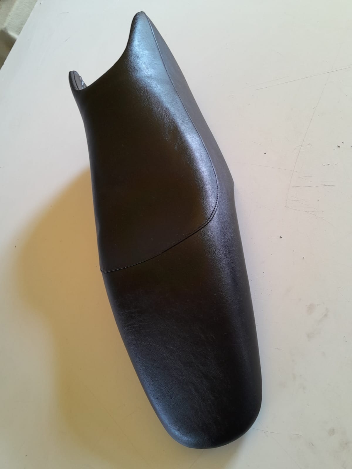 Czarne, skórzane obicie siedzenia motocyklowego, leżące na jasnym stole. Widoczny szew łączący dwie części materiału.