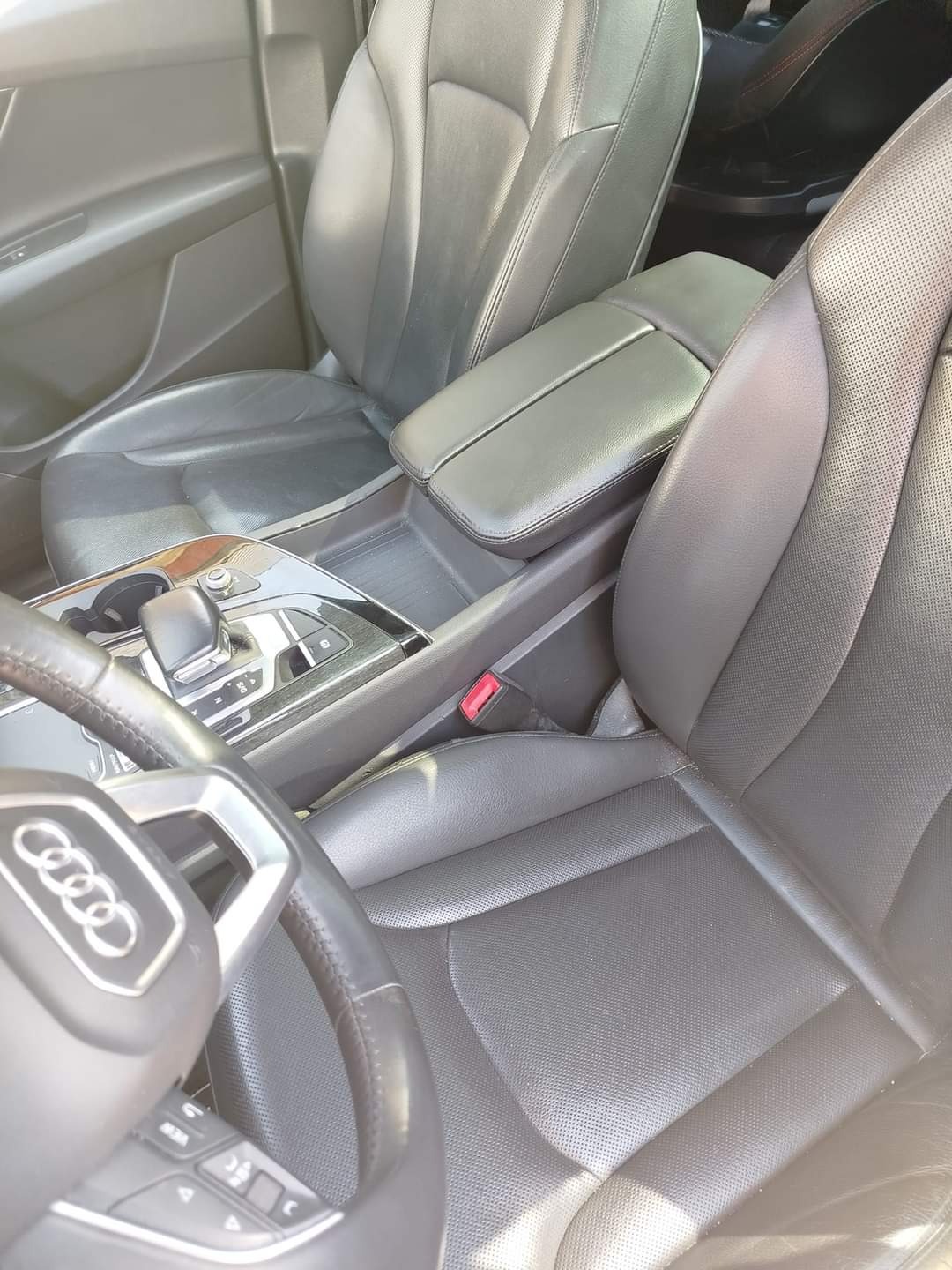 Wnętrze samochodu Audi Q7 z czarną skórzaną tapicerką foteli, widoczny fragment kierownicy z logo Audi, konsola środkowa i fotel pasażera, skóra miejscami lekko zużyta.