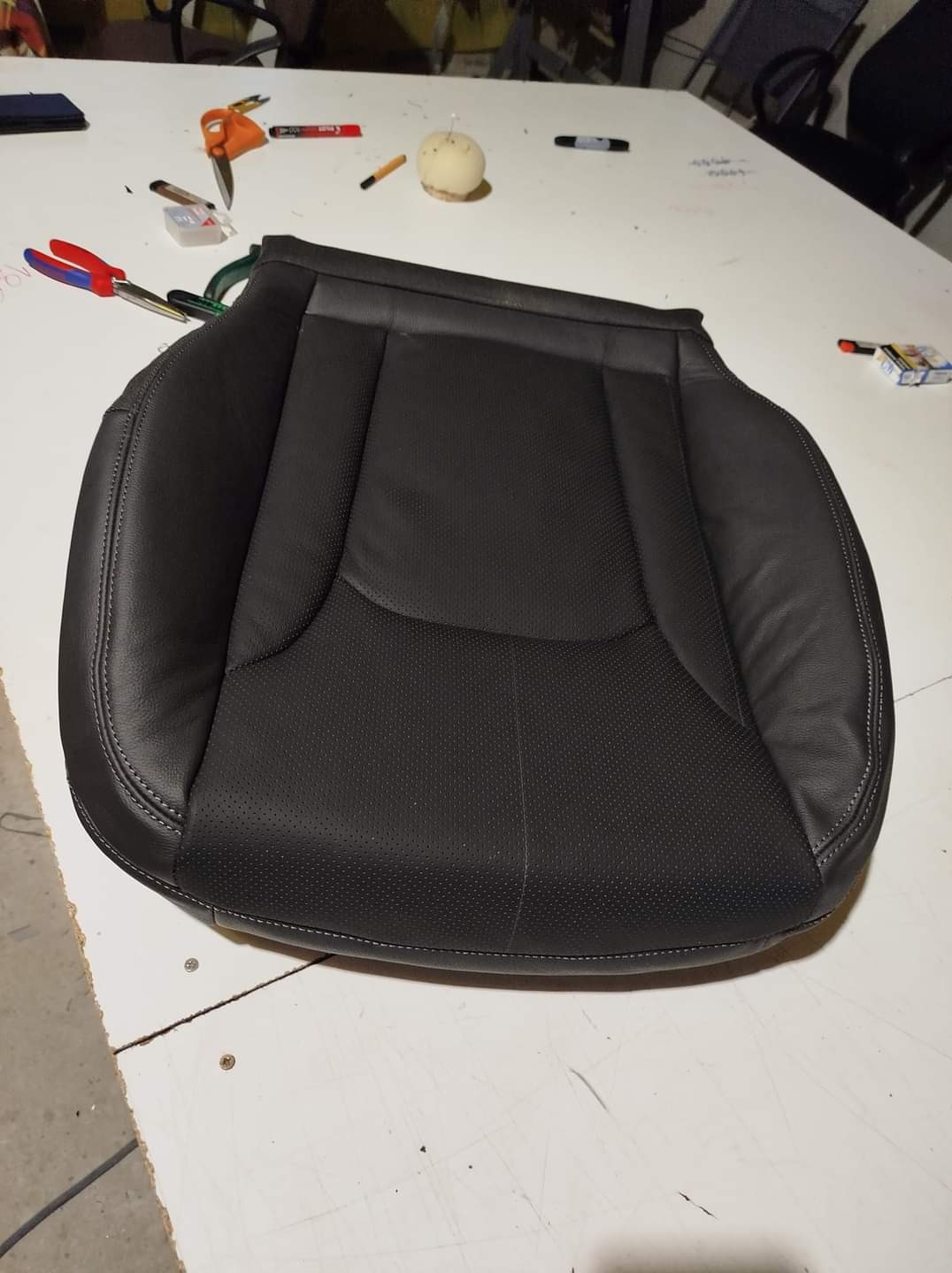 Czarne, skórzane siedzisko samochodowe z perforowaną powierzchnią, leżące na białym stole warsztatowym, obok narzędzi i akcesoriów.
