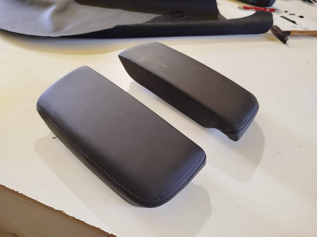 Dwa czarne, tapicerowane podłokietniki na białym stole warsztatowym, widoczne narzędzia i materiały tapicerskie w tle.