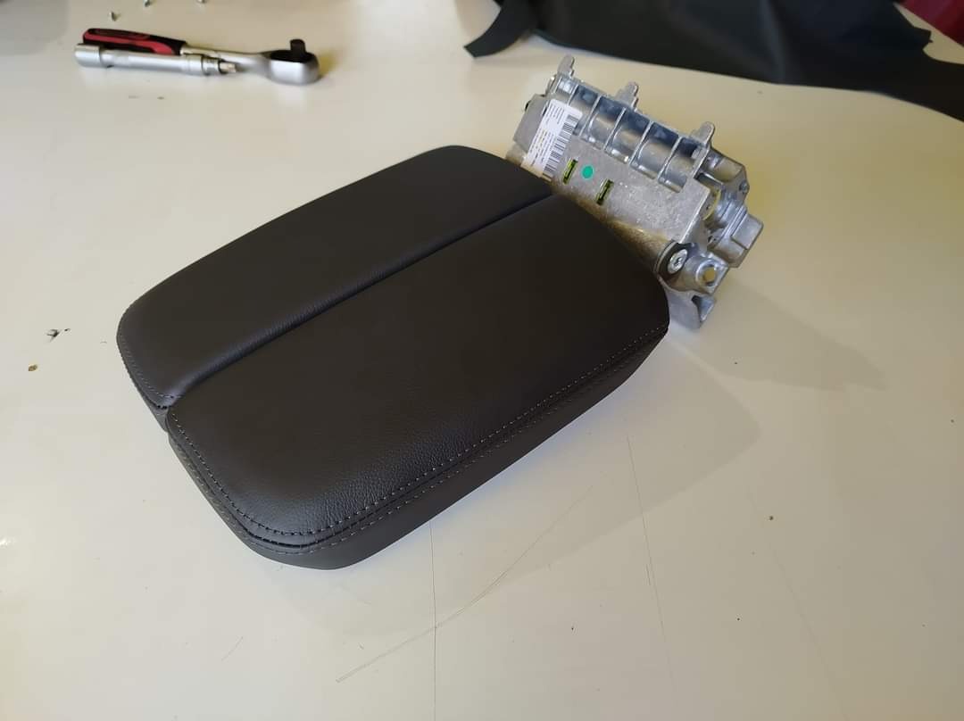 Ciemnoszary, skórzany podłokietnik samochodowy z widocznym mechanizmem mocującym i kluczem nasadowym w tle.