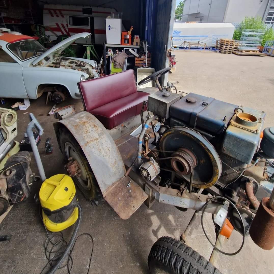 Renowacja starego traktora: czerwona tapicerka siedzenia na tle silnika i karoserii, obok odkurzacz, w tle biały samochód z otwartą maską w warsztacie.