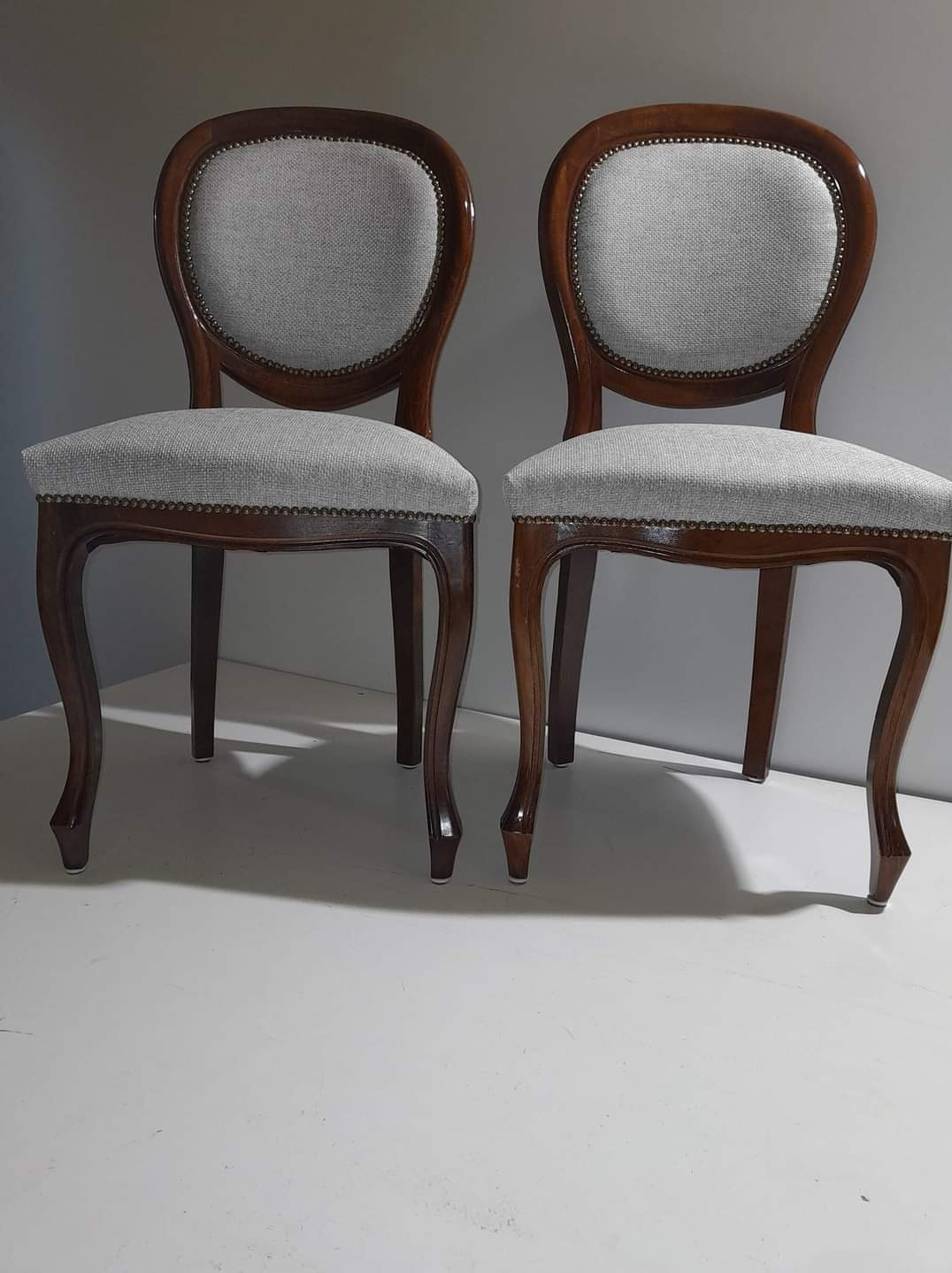 Dwa krzesła w stylu Ludwika Filipa z szarą tapicerką i ozdobnymi gwoździami, ustawione obok siebie na białym tle.