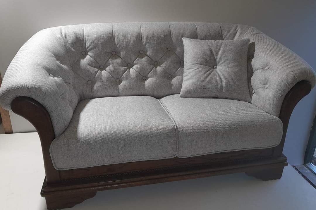 Dwuosobowa sofa z jasnoszarą tapicerką w stylu Chesterfield, z drewnianymi podłokietnikami i nogami, uzupełniona poduszką w tym samym odcieniu.