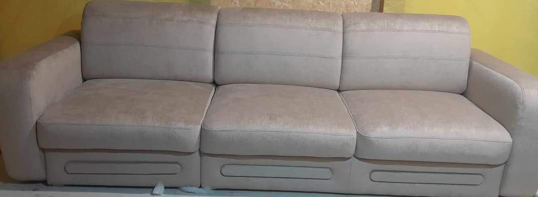 Beżowa, trzyosobowa sofa z welurową tapicerką, widoczne podłużne przeszycia na oparciu i siedzisku, dolna część z dekoracyjnymi wgłębieniami.