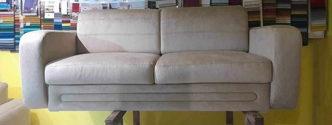 Jasnoszara, dwuosobowa sofa z delikatnymi przeszyciami, ustawiona na drewnianej konstrukcji, z widocznymi próbkami tkanin w tle.