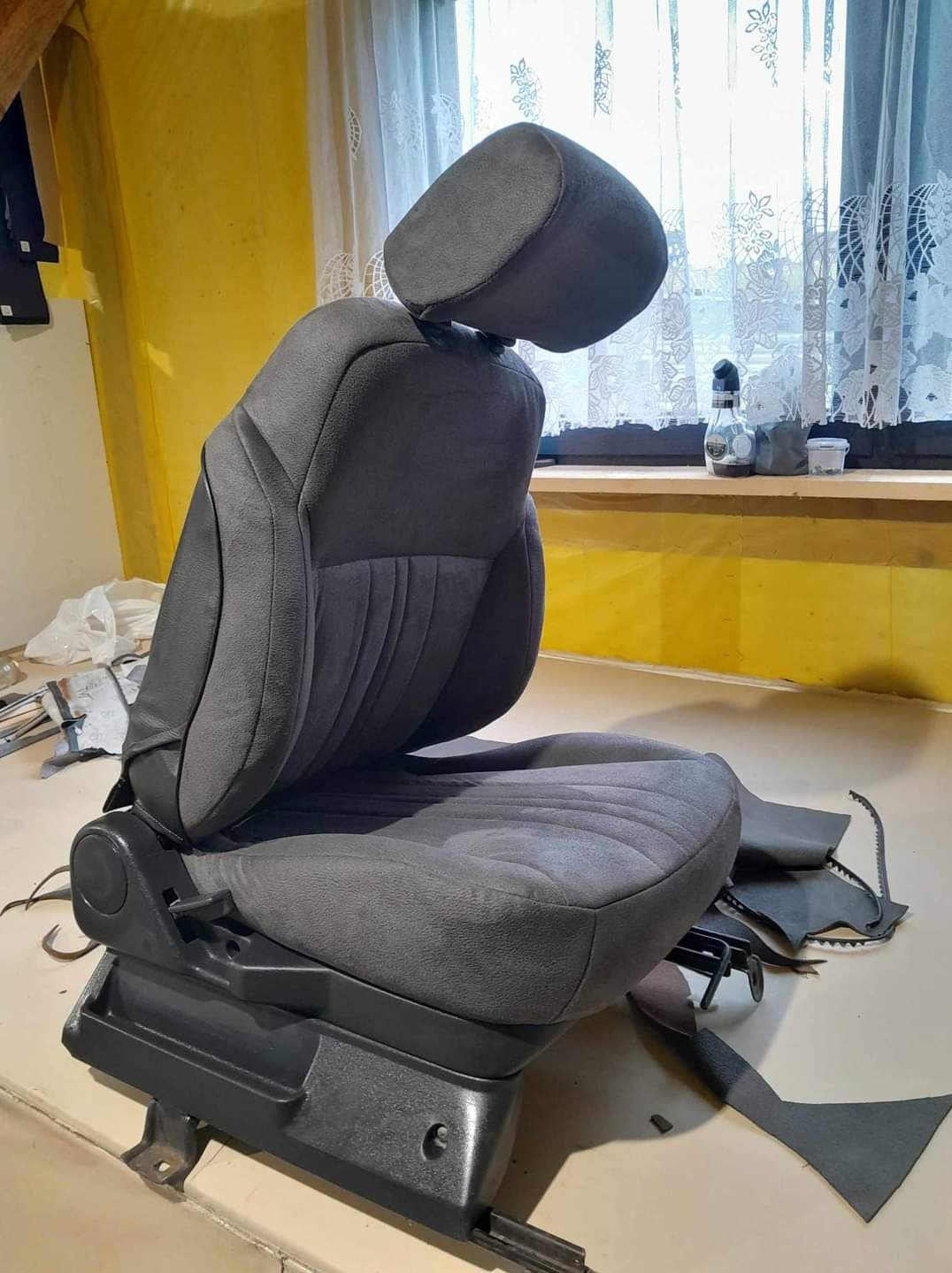 Szare samochodowe siedzenie z zagłówkiem, częściowo obite nowym materiałem, widoczne narzędzia i fragmenty starej tapicerki na stole.