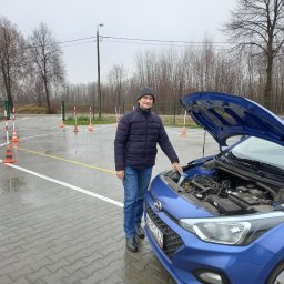 OSKK PEGAZ Zbigniew Muszyński - Instruktor obok niebieskiego samochodu z otwartą maską na placu manewrowym, widoczne pachołki i białe linie wyznaczające tor jazdy.