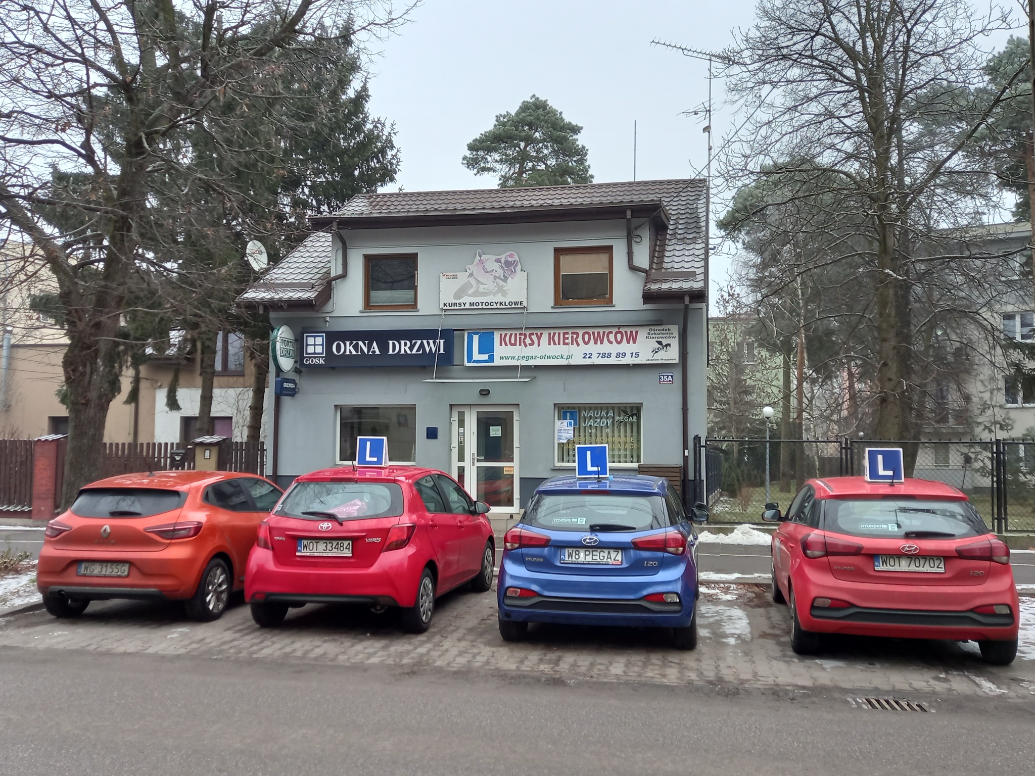 Budynek z szyldami 'Okna Drzwi', 'Kursy Motocyklowe', 'Kursy Kierowców', przed którym zaparkowane są cztery samochody z literą 'L' na dachu.