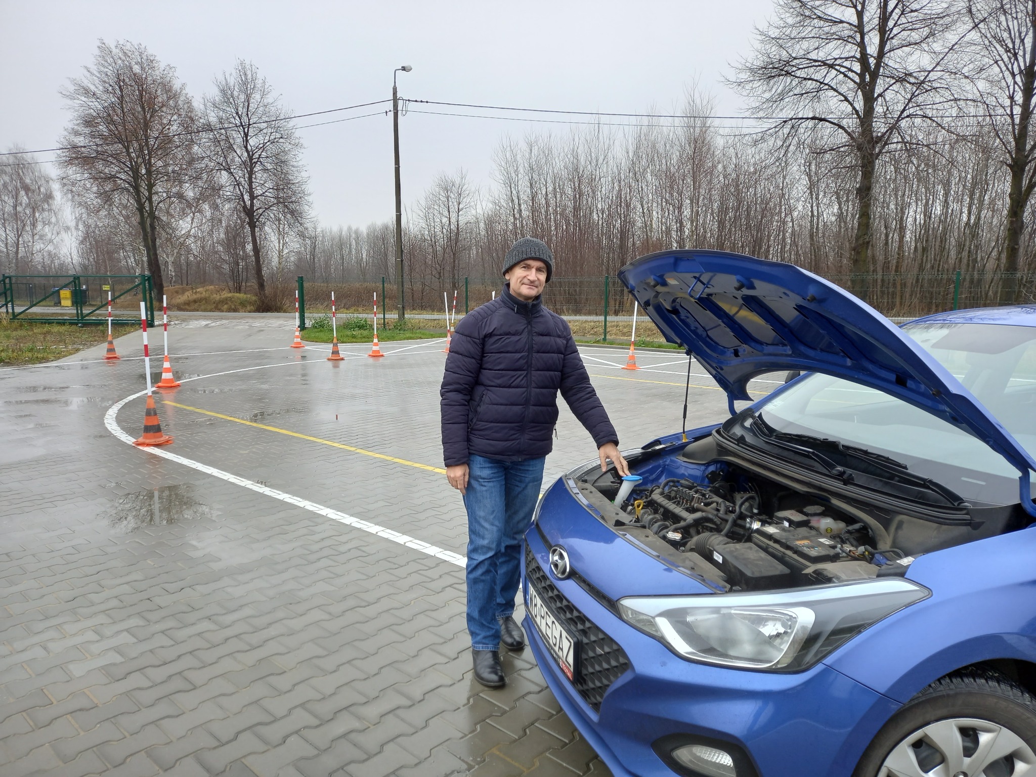 Instruktor obok niebieskiego samochodu z otwartą maską na placu manewrowym, widoczne pachołki i białe linie wyznaczające tor jazdy.