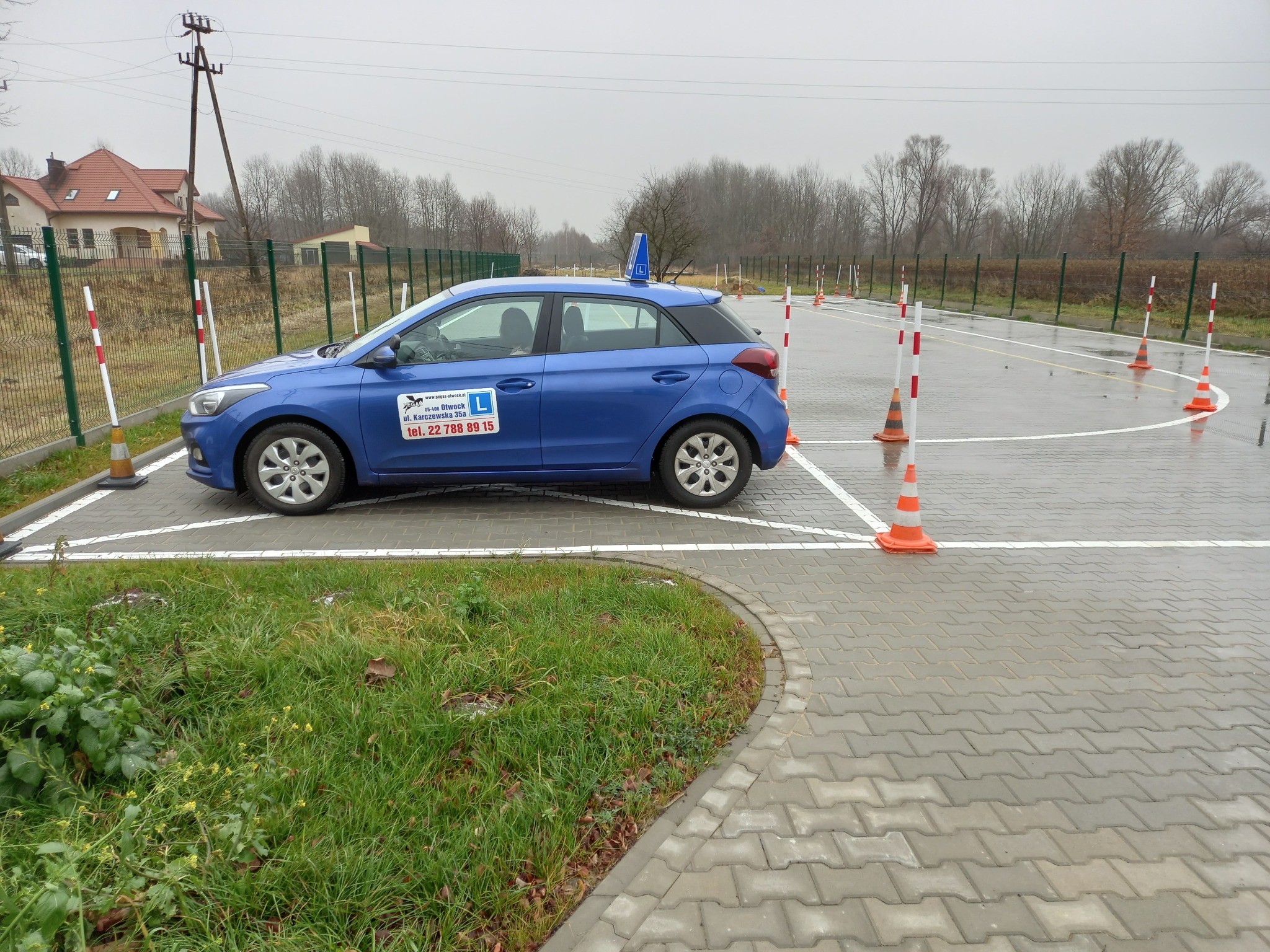 Niebieski samochód z literą L na dachu, zaparkowany na placu manewrowym z pachołkami i liniami, widoczne logo na drzwiach pojazdu, mokra nawierzchnia, pochmurny dzień.