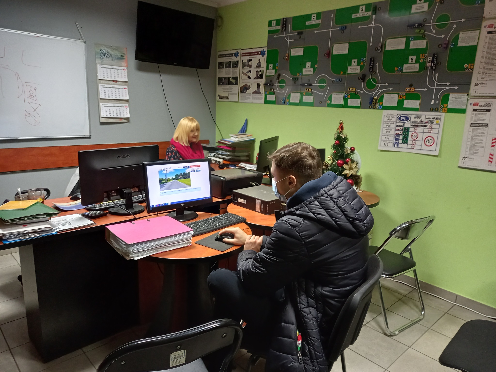 Biuro z pracownikiem w maseczce przy komputerze, instrukcje i schematy na ścianie, kalendarze, monitor z symulacją jazdy, dekoracja świąteczna na biurku.