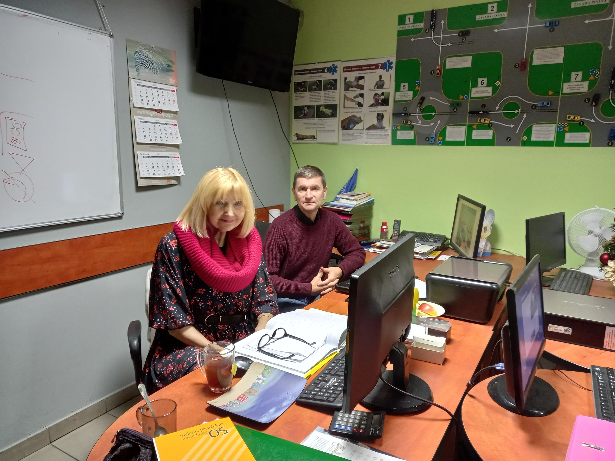 Instruktorzy w biurze, na ścianie schemat toru egzaminacyjnego, kalendarze i instrukcje pierwszej pomocy.