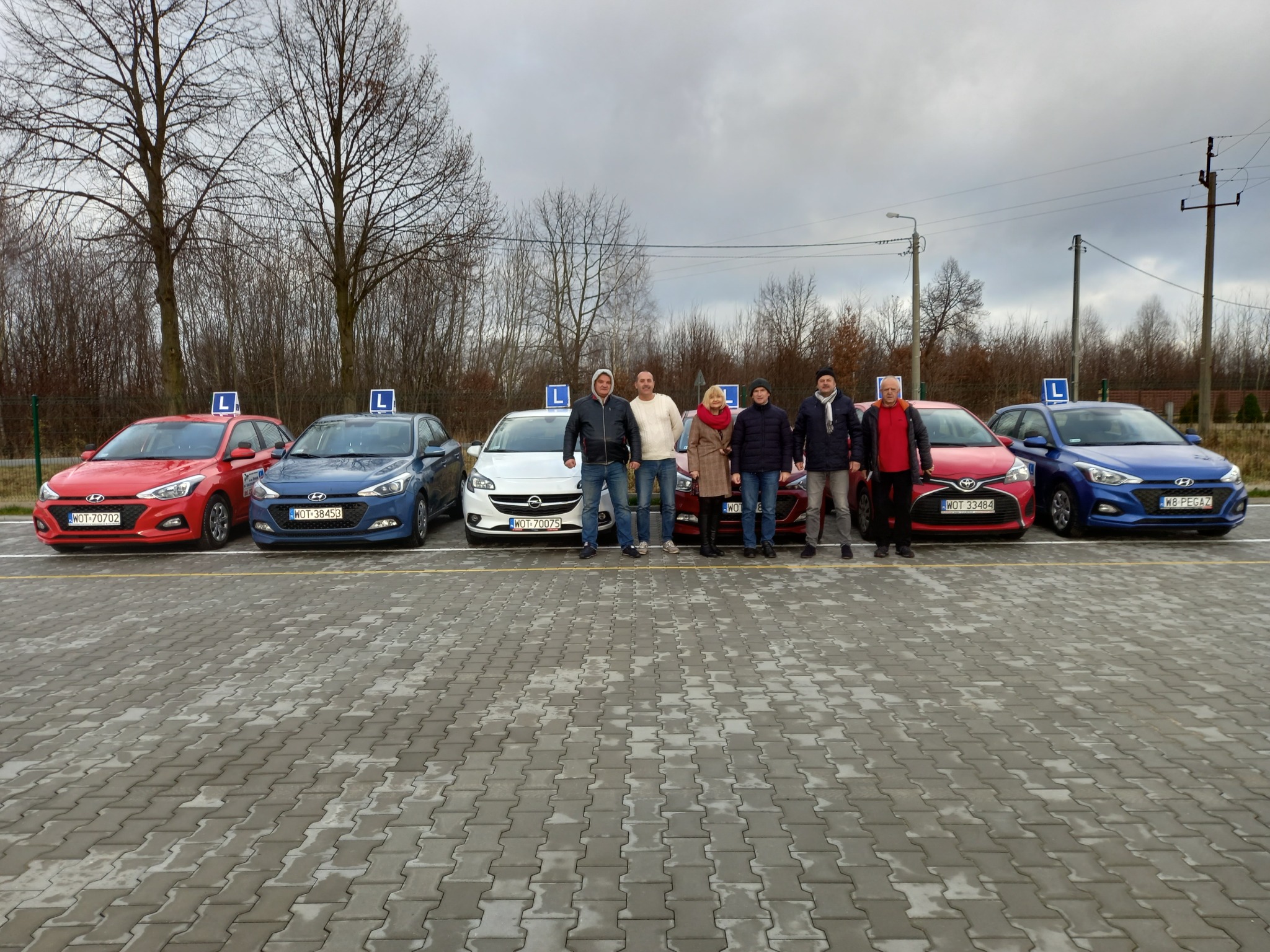 Grupa osób stoi przed samochodami oznaczonymi literą L na dachu, zaparkowanymi na placu z kostki brukowej w pochmurny dzień.