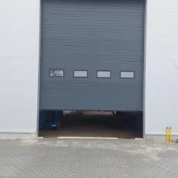 Częściowo otwarta, szara brama segmentowa z czterema okienkami, zamontowana w białej hali przemysłowej. Widoczny fragment podnośnika wewnątrz.