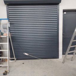 iTiro Jakub Zieliński - Zamknięta, szara brama rolowana z widocznymi drabinami monterskimi w Wielogłowach. Widoczny kabel zasilający i gniazdko elektryczne na podłodze.