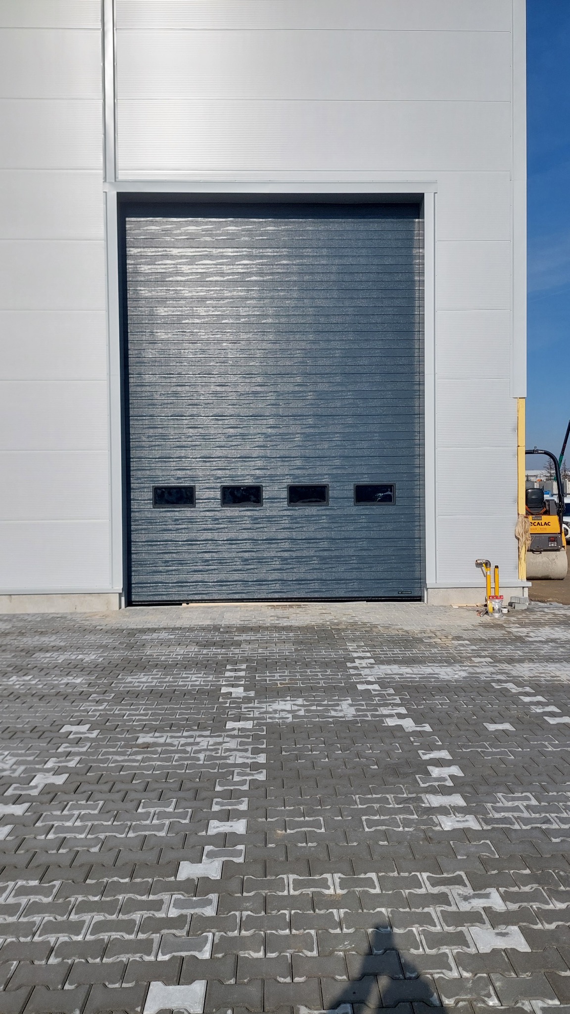 Szara brama przemysłowa segmentowa z czterema małymi oknami, widoczna na tle białej ściany hali i częściowo wybrukowanego placu z kostki.