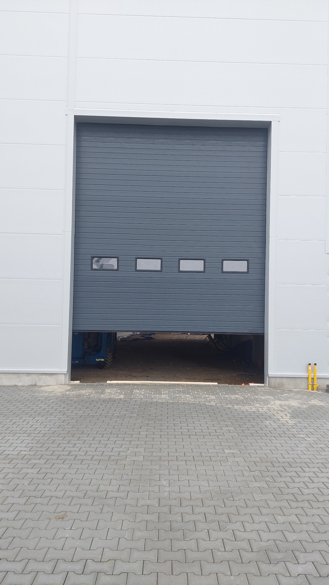 Częściowo otwarta, szara brama segmentowa z czterema okienkami, zamontowana w białej hali przemysłowej. Widoczny fragment podnośnika wewnątrz.