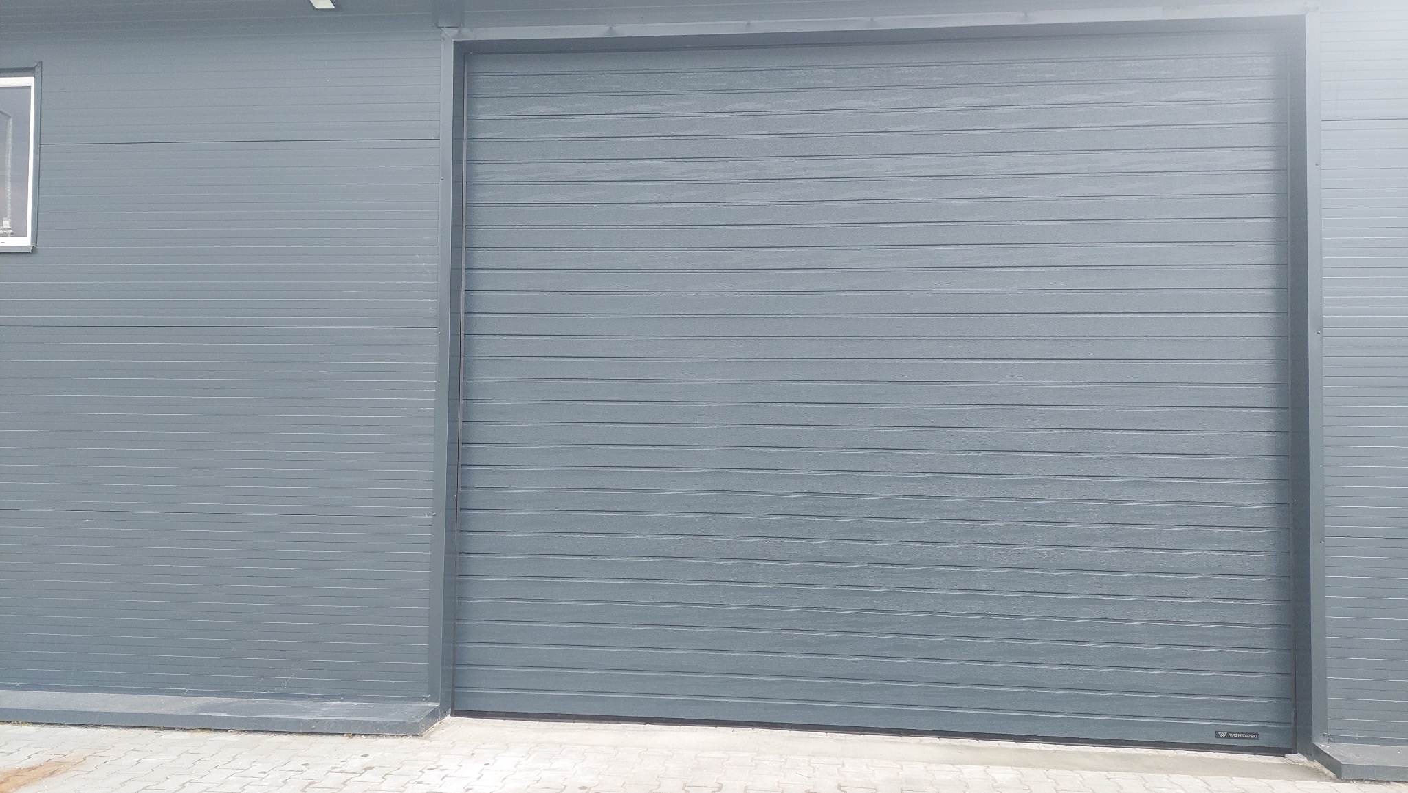 Szara brama segmentowa garażowa, zamknięta, z poziomymi panelami. Widoczny fragment elewacji budynku z oknem. Niewielki fragment kostki brukowej u dołu.